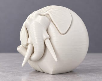 May include: A spherical off-white sculpture featuring a detailed elephant head and trunk integrated into the rounded form. The surface displays fine horizontal lines and textures, with the elephant's ears, tusks, and trunk protruding from the side.