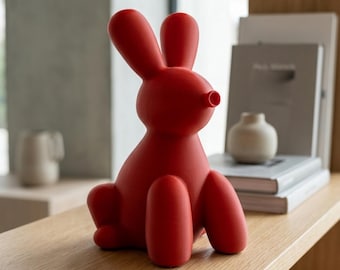 May include: A matte red rabbit figurine with a smooth, rounded, and stylized form. The rabbit is depicted in a seated position with long, upright ears and a small, circular snout. The surface of the object has a uniform, solid red color.