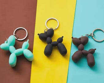 May include: Three balloon dog keychains in mint green, matte black, and dark brown. Each keychain features a silver-toned metal split ring attached to a smooth, molded figure that mimics the appearance of a twisted balloon animal dog shape.