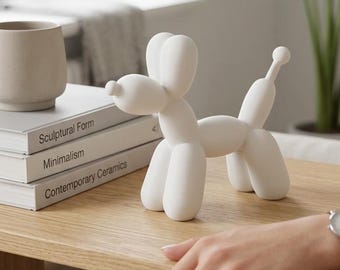 May include: A matte white ceramic sculpture shaped like a balloon dog, featuring rounded limbs, a long snout, and a small tail. The sculpture is positioned on a wooden surface next to a stack of three books titled Sculptural Form, Minimalism, and Contemporary Ceramics.