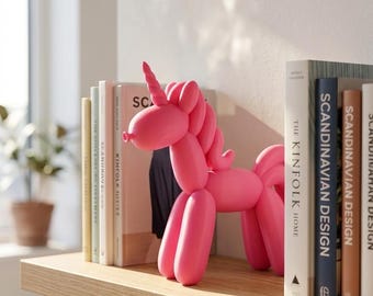 May include: A bright pink sculpture shaped like a balloon animal unicorn, featuring a smooth, matte finish, a pointed horn, a rounded snout, and a stylized mane and tail, standing upright on a flat surface between several hardcover books.