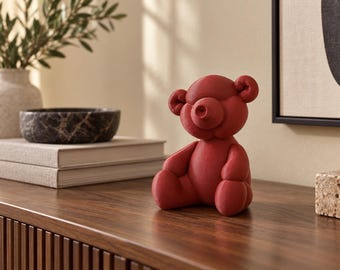 May include: A matte red sculpture of a seated teddy bear with a smooth, rounded form. The bear features a prominent circular snout, small rounded ears, and a simple, stylized body shape. The figure is rendered in a solid, deep red color.