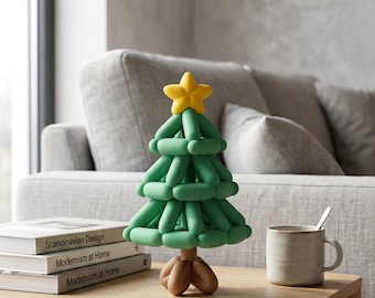 May include: A decorative Christmas tree sculpture constructed from green, brown, and yellow balloon-like shapes. The tree features a yellow star topper, green branches arranged in tiers, and a brown base, standing on a wooden table next to books.