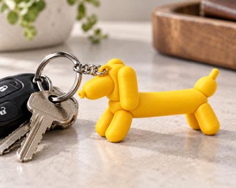 May include: A bright yellow keychain shaped like a balloon animal dog, attached to a silver-toned metal split ring holding two standard house keys and a black car key fob. The dog figure has a smooth, matte finish and a simple, stylized form.