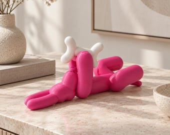 May include: A bright pink sculpture shaped like a balloon animal dog, featuring a smooth, rounded form. The dog is depicted in a lying position, holding a small, solid white bone-shaped object between its front paws, crafted with a glossy finish.