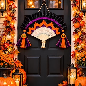 May include: A decorative wall hanging shaped like a folding fan, featuring a black, purple, and orange chevron pattern. The fan includes two dangling tassels with orange jack-o'-lantern beads, suspended by a rope on a black door.