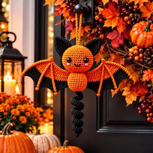 Op de afbeelding: Een gehaakt vleermuisornament met een ronde oranje kop met zwarte oren, een klein oranje lichaam en brede vleugels die aan de bovenkant oranje en aan de onderkant zwart zijn. Een zwarte spiraalvormige staart hangt onderaan, met een oranje koord.