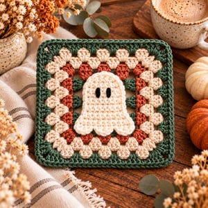 May include: A square crochet granny square coaster featuring a central cream-colored ghost with black embroidered eyes. The square is bordered by concentric layers of cream, rust-orange, and dark forest green yarn in a classic cluster stitch pattern.