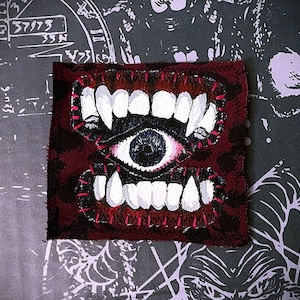May include: A square fabric patch featuring a hand-painted design of a central human-like eye surrounded by upper and lower rows of sharp white teeth. The background is a dark mottled red, and the edges are finished with visible pink stitching.