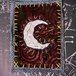 May include: A rectangular fabric patch featuring a dark brown and red paisley pattern background. A white crescent moon is embroidered in the center. The patch is bordered by a light blue denim edge with bright yellow cross-stitch embroidery.