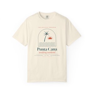 May include: A cream-colored short-sleeve t-shirt featuring a graphic of a palm tree, sun, and ocean waves inside an arch. The text reads THE OFFICIAL PROGRAM, Punta Cana wedding weekend, FRI: WELCOME DRINKS, SAT: I DO'S, SUN: RECOVERY BRUNCH.
