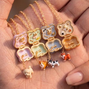 Pode incluir: Quatro colares com medalhão em metal dourado com estojos articulados e interiores em esmalte pastel. Cada medalhão contém pequenos berloques em forma de cão, incluindo um caniche, um corgi, um bulldog francês e um Shiba Inu.