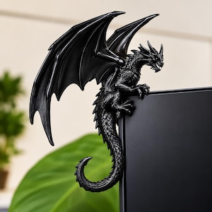 May include: A black dragon figurine with detailed scales and large, spread wings. The creature is perched on the top corner of a flat, rectangular black screen, with its long, curved tail hanging down the side of the frame in a static pose.