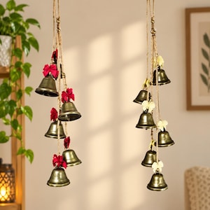 May include: Two vertical hanging decorations featuring multiple brass-colored metal bells suspended on natural jute twine. The left strand is adorned with bright red fabric bows, while the right strand features pale yellow fabric bows.