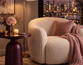 May include: A cream-colored, textured bouclé armchair featuring a rounded back and seat. The chair is adorned with a dusty rose velvet throw pillow and a matching fringed throw blanket. A dark purple pedestal side table sits beside the chair.