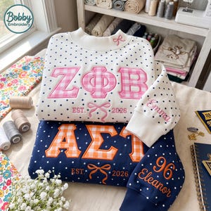 May include: A white sweatshirt with black polka dots features pink gingham Greek letters, a bow, and the text "EST. 2026" with "Emma" on the sleeve. Below, a navy sweatshirt with white polka dots has orange gingham Greek letters and "Eleanor 96".