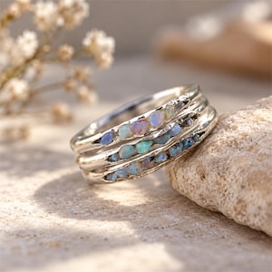 May include: A silver ring featuring three stacked bands, each embedded with a row of small, raw, multicolored opal stones displaying iridescent flashes of blue, pink, and green against the metallic surface of the jewelry piece.