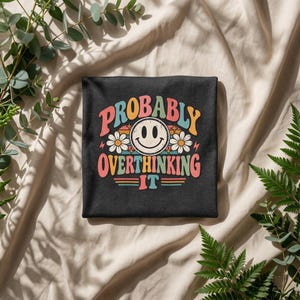 May include: A dark gray t-shirt featuring a graphic print with the text "PROBABLY OVERTHINKING IT" in a multi-colored retro font. The design includes a central smiley face, two white daisies, small star accents, and horizontal lines.