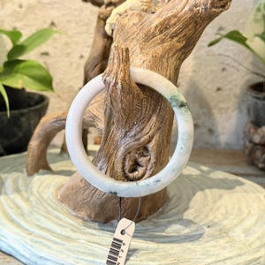 May include: A smooth, circular bangle bracelet crafted from pale, translucent stone. The bangle features subtle, mottled green inclusions distributed throughout its polished surface, and it includes a small white paper tag attached by a thin string.
