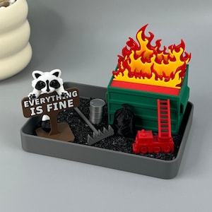 May include: A miniature desk display featuring a green dumpster engulfed in red and yellow flames, a raccoon figurine holding a brown sign that reads EVERYTHING IS FINE, a small red fire truck, a black trash bag, a silver barrel, and a rake.