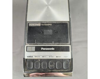 Può includere: Un registratore a cassette portatile Panasonic nero e argento con altoparlante montato in alto, vano centrale per cassette e pannello di controllo con sei pulsanti neri etichettati Record, Rewind, FF, Play, Stop ed Eject.