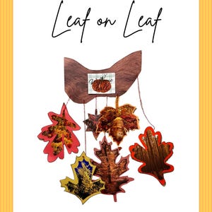 Op de afbeelding: Een decoratieve wandhanger met de tekst Leaf on Leaf bovenaan. Daaronder toont een houten basis een klein vierkant bordje met de tekst Welcome Fall. Vijf bladvormige uitsparingen met diverse herfstbosafbeeldingen hangen aan de basis.