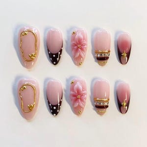 Puede incluir: Un juego de diez uñas postizas en forma de almendra con base rosa claro. Los diseños incluyen marcos de alambre dorado, puntas marrón oscuro con lunares blancos, flores rosas en relieve, bandas doradas horizontales con pedrería y dijes de estrellas doradas.