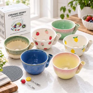 May include: A collection of ceramic berry colander bowls in various colours and patterns, including solid blue, green, yellow-pink gradient, and white with strawberry or lemon prints. Each bowl features a side handle and drainage holes.