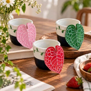 Può includere: Tre tazze in ceramica con una sezione superiore bianca e una inferiore blu scuro, ciascuna con un motivo a vite verde dipinto a mano. Sul lato di ogni tazza è presente un grande manico a forma di foglia testurizzata rosa, verde o rossa.