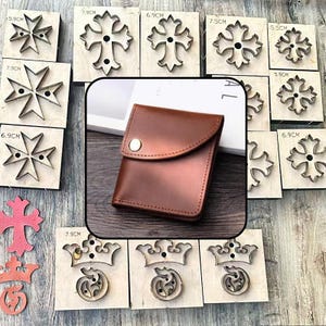 May include: A brown leather wallet with a single silver snap button closure is centered among various metal cross and crown-shaped cutting dies mounted on wooden blocks, each labeled with metric measurements like 5.9cm and 7.9cm.