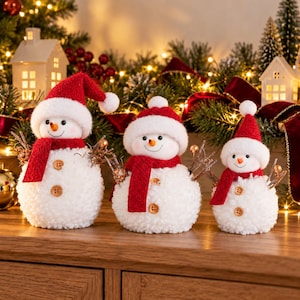 May include: Three white plush snowman figurines of varying sizes, each wearing a red knit hat with a white pom-pom and a matching red scarf. Each snowman features two brown wooden buttons on the front and twig-like arms with small gold accents.