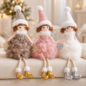 May include: Three plush dolls with brown hair, wearing knit hats with white pom-poms and faux fur trim. The dolls have fluffy skirts, dangling legs with white fur cuffs, and metallic boots. They hold a gold star, a gold tree, and a silver heart.