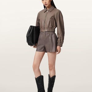 May include: A taupe leather jacket with a front zipper, collar, and elasticized hem and cuffs, paired with matching taupe leather shorts featuring an elastic waistband. A black leather handbag and black leather cowboy boots complete the outfit.