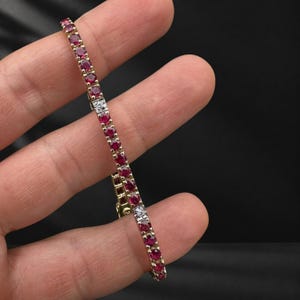 May include: A gold tennis bracelet featuring a continuous row of round, deep red rubies set in metal prongs, interspersed with two small, square-shaped silver-toned links containing tiny, clear, sparkling accent stones.