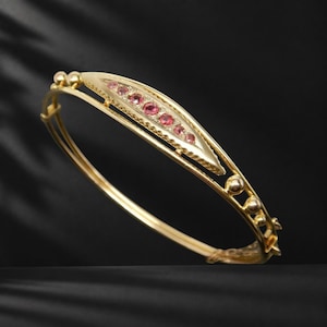 May include: A gold bangle bracelet featuring a central elongated, pointed element set with five small, round, pink gemstones. The band is constructed from thin, polished gold wire with small gold spheres spaced along the sides.