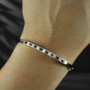 May include: A polished metal bangle bracelet featuring a channel-set row of alternating square-cut deep red rubies and clear white diamonds. The slim, rigid band has a hinged closure and a smooth, reflective finish along the outer surface.