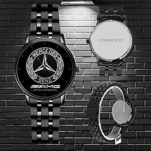 Pode incluir: Um relógio de pulso preto com pulseira de elos metálicos. O mostrador apresenta o logótipo da Mercedes-Benz e o texto AMG DRIVING PERFORMANCE. Uma vista mostra a parte de trás da caixa com o texto CUSTOM TEXT e outra o perfil lateral.