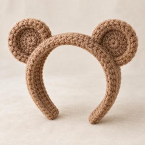 May include: A light brown crochet headband featuring two circular, plush, bear-like ears attached to the top. The entire accessory is crafted from a single-colored yarn with a visible, textured stitch pattern throughout the band and the rounded ears.