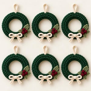 May include: Six circular crochet ornaments in dark green, each featuring a small cream-coloured hanging loop at the top, a cream-coloured bow at the bottom, and a cluster of three dark red berries with two light green leaves on the side.