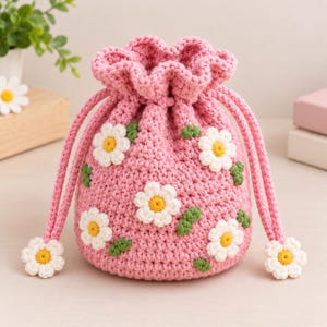 Puede incluir: Una bolsa de ganchillo rosa con cordón ajustable, decorada con aplicaciones de margaritas blancas con centros amarillos y pequeñas hojas verdes. El bolso tiene un borde superior con volantes y dos largos cordones rosas con margaritas.