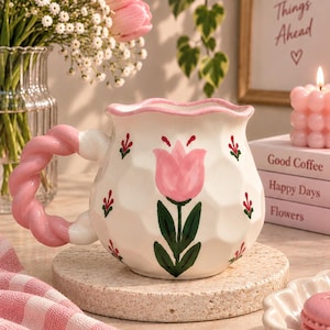 May include: A white ceramic mug with a scalloped rim and a twisted pink handle. The mug features a hand-painted pink tulip with green leaves on the front, surrounded by small red floral accents. The surface of the mug has a textured, dimpled finish.