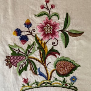 May include: A piece of off-white fabric featuring a colorful floral embroidery design. The pattern includes a large pink flower, green leaves, yellow accents, and small blue berries, all stitched with detailed thread work on a plain textile base.