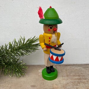 May include: A wooden figurine of a drummer in a yellow jacket and green hat, holding drumsticks and a drum. The drum is blue, red, and white. The figurine has a red nose and a red feather in its hat. The figurine is on a green base.
