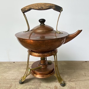 May include: Antique copper tea kettle with a wooden handle and brass accents. The kettle sits on a matching copper and brass stand with a burner. The kettle has a round body, a spout, and a lid with a black knob. A vintage kitchenware item.