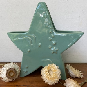 May include: A teal star-shaped ceramic vase with embossed star and swirl designs. The vase is displayed on a wooden surface with dried white flowers. The vase is a decorative item for home decor.