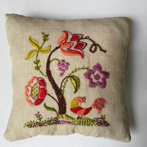 May include: A small, beige linen pillow with colorful floral embroidery. The embroidery features a tree with red, pink, and purple flowers, green leaves, and a small bird. The pillow is square and appears to be hand-stitched.
