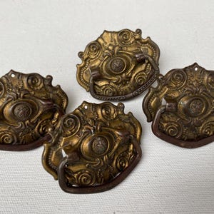 May include: Four antique brass drawer pulls with ornate, swirling designs. Each pull features a central medallion and a curved handle. The pulls are aged with a warm, golden hue, suggesting vintage craftsmanship. Ideal for furniture restoration projects.