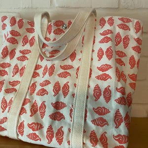 May include: A white tote bag with a repeating pattern of red croissant shapes. The bag has cream-colored straps and a snap closure. The bag is made of a sturdy fabric, perfect for carrying groceries or other items.