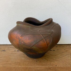 May include: A rounded ceramic vase with a wide, undulating rim and a tapered base. The surface features a metallic, iridescent finish in shades of copper, bronze, and dark brown, with a raised, abstract linear design embossed on the side.