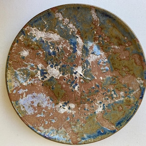 May include: A round ceramic plate with a unique, textured surface. The plate features a blend of colors, including brown, blue, green, and white, creating a marbled effect. The rim is a darker shade of blue. This decorative plate is suitable for serving food.